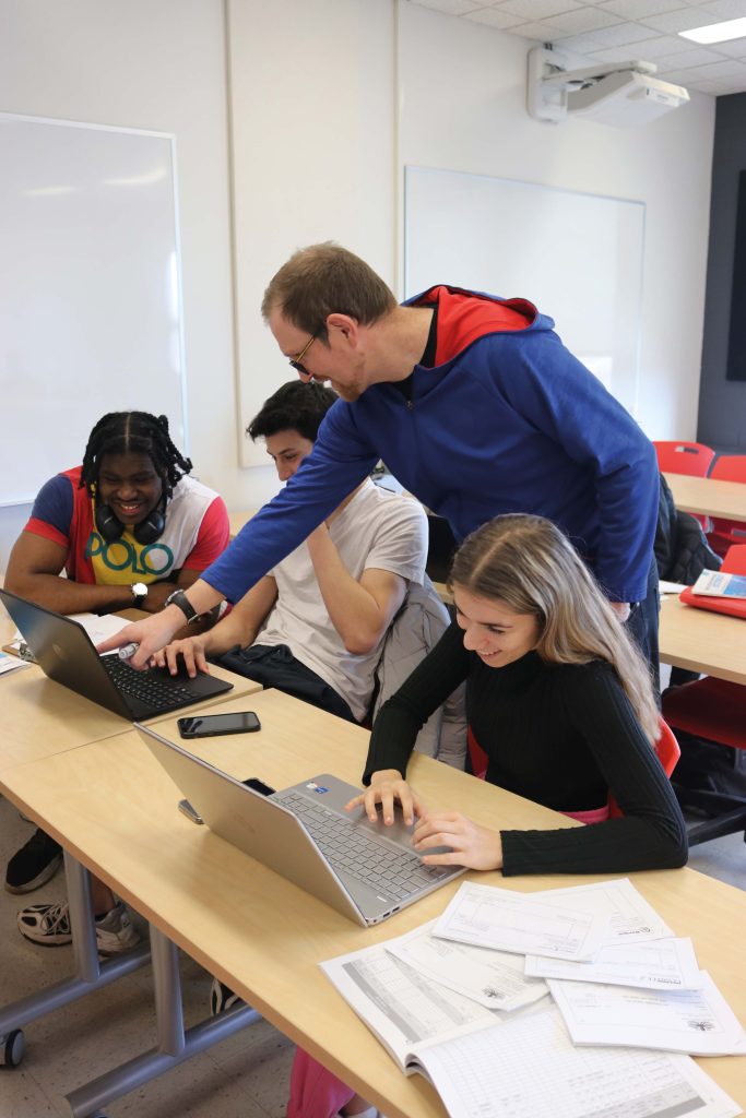 3 étudiants et 1 professeur dans une salle de classe, travaillant sur l'ordinateur. Le professeur pointe l'écran de l'ordinateur.