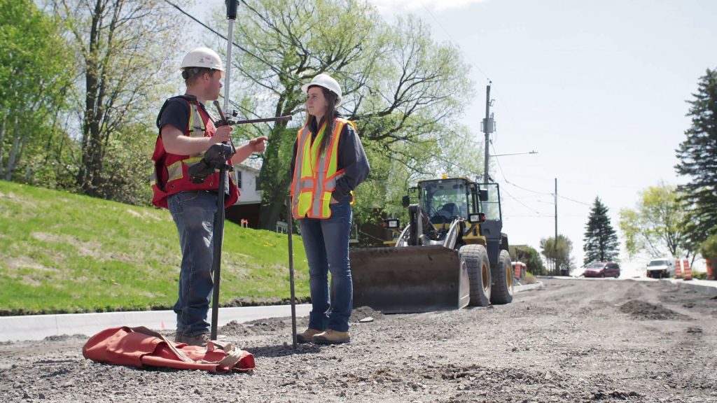 Deux étudiants face-à-face avec un dossard jaune et un casque de construction blanc sur une route résidentielle en construction.