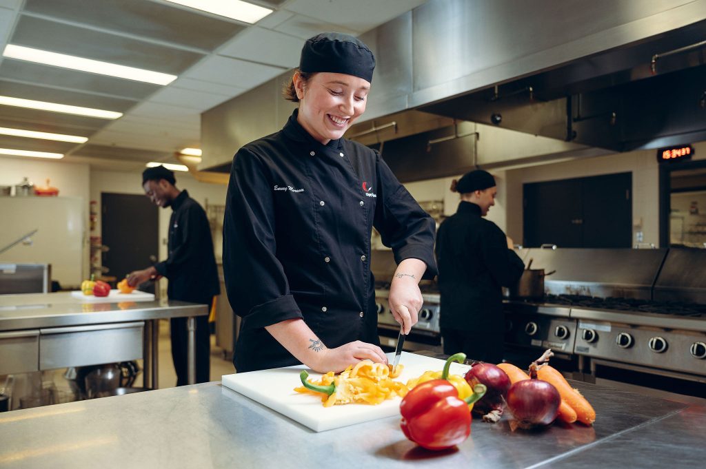 Étudiante en habit de cuisinière coupant des poivrons dans une cuisine académique.