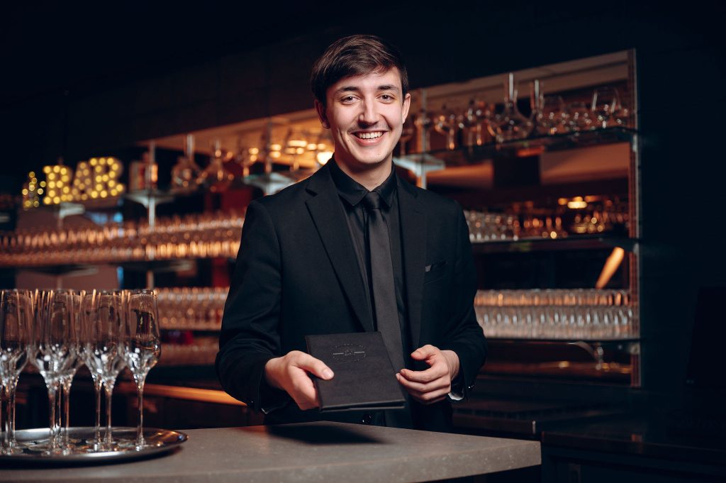 Étudiant en tuxedo dans le bar d'un hôtel, tendant un livret de menu.