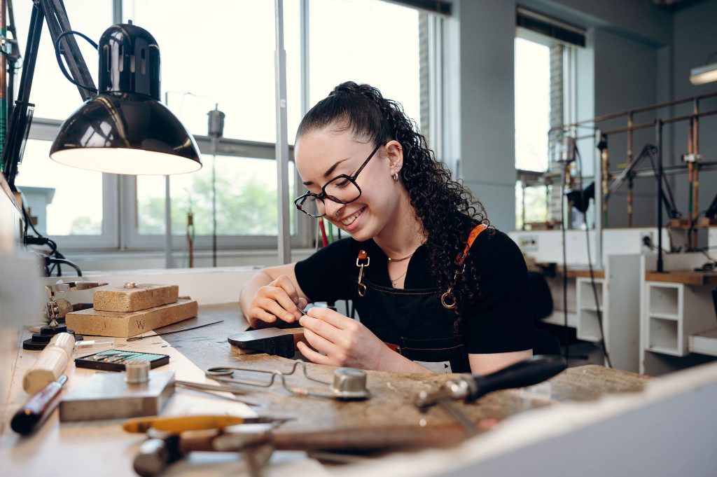 Étudiante dans un atelier, avec un tablier, penchée sur un bureau de travail et manipulant des outils de joaillerie.