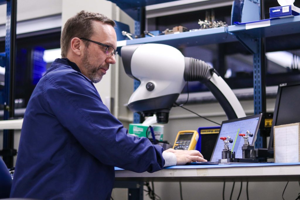 Homme habillé en bleu dans un atelier, qui regarde un tableau sur un ordinateur portable.
