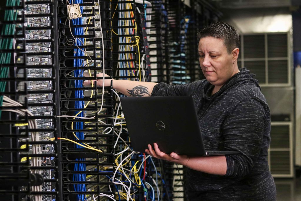 Femme devant un mur de fils réseau, regardant un ordinateur tout en manipulant les fils.