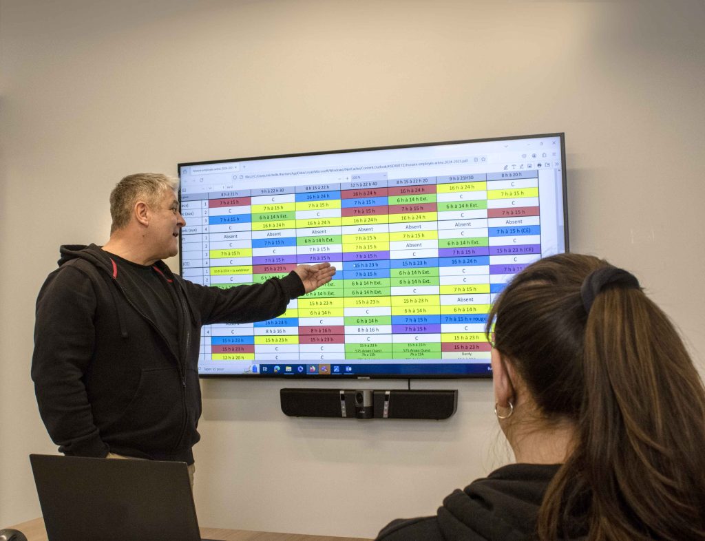 Homme devant un écran de télévision qui pointe un tableau affichant un horaire. Une femme regarde.