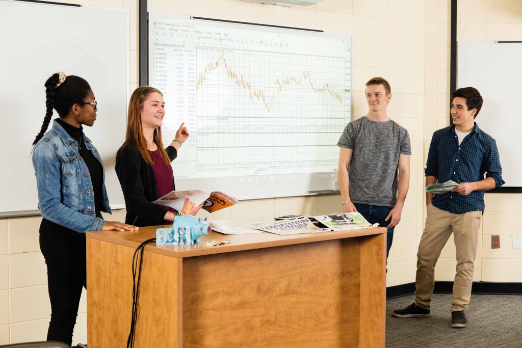 Groupe de quatre étudiants faisant une présentation dans une classe, pointant la projection d'un tableau d'économie.