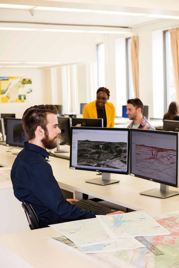 Étudiant dans un laboratoire informatique avec des cartes sur son bureau et regardant des images de cartographie sur son écran.