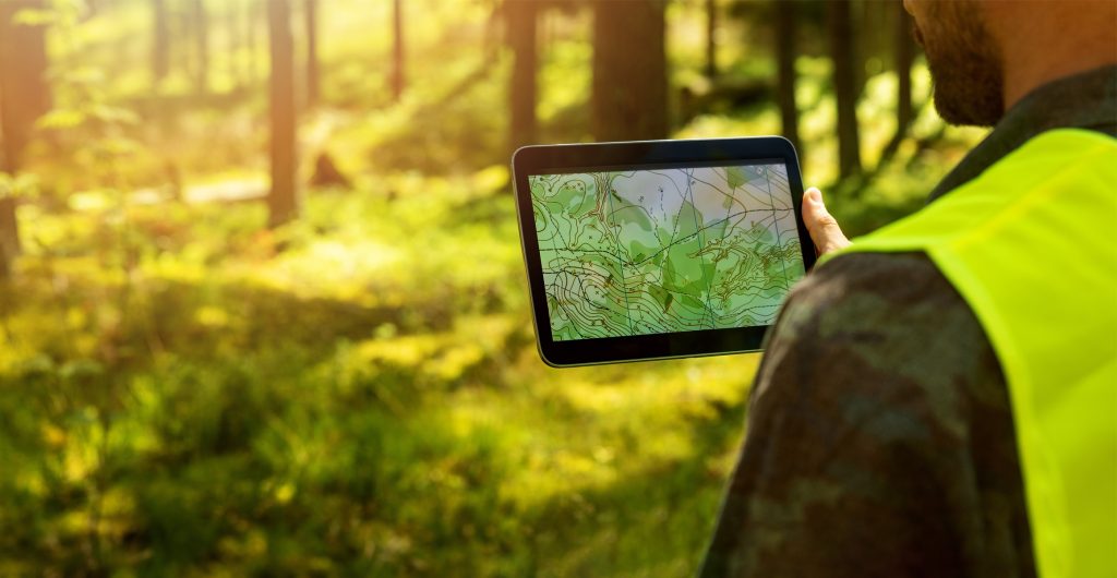 Homme avec un dossard jaune dans une forêt, regardant le plan du terrain sur une tablette.