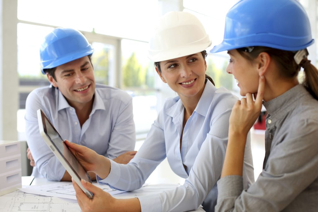 Deux femmes et un homme portant un casque de sécurité et regardant des plans architecturaux sur papier et sur tablette.